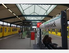 Berliner U-Bahnhof Gleisdreieck mit einem gelben U-Bahn-Zug auf der linken Seite, einem überdachten Glasdach mit Stahlkonstruktion und grünen Treppengeländern in der Mitte. Im Vordergrund sind ein Stationsschild und Sitzbänke mit wartenden Fahrgästen zu sehen.