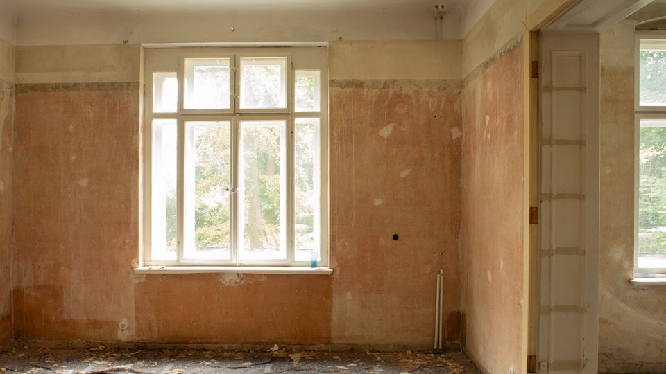 Innenraum eines leerstehenden, renovierungsbedürftigen Zimmers mit großem, weiß gerahmtem Fenster und abgenutzten, teils beschädigten Putzwänden. Rechts führt ein breiter Durchgang in einen weiteren Raum mit zusätzlichem Fenster; auf dem Boden liegen Schutt und abblätternde Materialien.