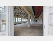 Großer, leerer Innenraum eines Gebäudes im Rohbauzustand mit sichtbarer Deckenstruktur aus freiliegenden Trägern, mehreren weißen Säulen und einer Reihe rechteckiger Fenster an den Wänden. Die Türrahmen im Vordergrund führen in den Raum, der noch unfertig ist.