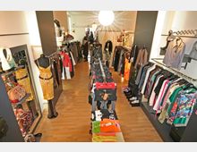 Innenansicht eines kleinen Modegeschäfts mit Holzfußboden, auf dem Kleiderständer, Regale mit Kleidung, Taschen und Accessoires ausgestellt sind. Zentral im Raum steht ein Tisch mit gefalteten Kleidungsstücken, während die Wände mit hängender Kleidung und Regalen dekoriert sind.