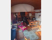 Wohnraum mit offenem Grundriss, Holzdach mit Rundbogen, gemusterten Teppichen und Holzböden. Der Raum umfasst eine Sitzecke, einen Esstisch mit roter Tischdecke, eine Küchenzeile mit Barhockern und Regale mit dekorativen Gegenständen.