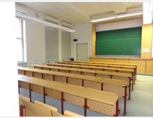 Ein leerer Hörsaal mit mehreren Reihen aus hellen Holzsitzbänken und Tischen, einer großen grünen Tafel an der Wand und Fenstern mit heruntergelassenen Rollläden auf der linken Seite. Die Decke ist flach mit Rasterleuchten ausgestattet, und der Raum hat eine nüchterne Einrichtung.