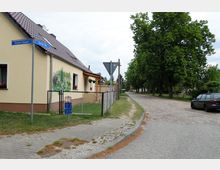 Straßenecke in einem Wohngebiet mit einem eingeschossigen Haus mit gelber Fassade und bemalter Wand, einem Hof mit Zaun und einem Straßenwegweiser zu den Straßen „Potsdamer Chaussee“ und „Berliner Weg“. Rechts verläuft eine alterschippe Kopfsteinpflasterstraße mit großen grünen Bäumen.