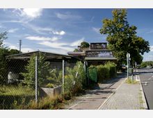 Außenansicht des Bahnhofs Wannsee, mit einem verwitterten Gebäude, auf dem Graffiti zu sehen ist, umgeben von einem Zaun und überwucherten Pflanzen. Rechts verläuft ein gepflasterter Gehweg an einer Straße mit Bäumen und Laternen.