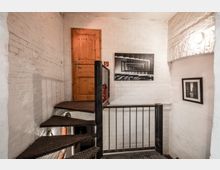 Industrieller Treppenhausbereich mit Metall-Wendeltreppe, weißen Ziegelwänden und einer Holztür im oberen Bereich; an der Wand hängen zwei Schwarz-Weiß-Fotografien, und ein Feuerlöscher befindet sich neben der Treppe.