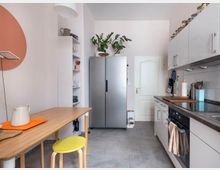 Moderne Küche mit hellen Wänden und grauem Fliesenboden. Zu sehen sind ein Esstisch aus Holz mit gelbem Hocker, ein großer silberner Kühlschrank, weiße Hängeschränke mit Vorratsgläsern sowie ein Herd und eine Spüle in eine dunkle Arbeitsplatte integriert.