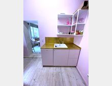 Kleine Küche mit rosa Wänden und Holzfußboden. Die Küchenzeile hat hellrosa Schränke, eine goldene Arbeitsplatte, ein weißes Spülbecken und weiße Regale mit Geschirr.