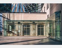 Moderne Glaskonstruktion mit dahinterliegenden historischen Fassadenelementen, die reich verzierte Fenster und dekorative Schnitzereien zeigen. Im Hintergrund sind das Dach des Sony Centers sowie Glas- und Stahlfassaden anderer Gebäude sichtbar.