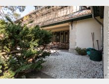 Ein gepflasterter Außenbereich mit Kiesboden und einer Terrasse, die teilweise von einem Dach mit roten Ziegeln überdacht ist. Im Vordergrund stehen Büsche, und im Hintergrund sind Metallstühle und ein Tisch unter einem verglasten Fenster mit Gittern zu sehen.