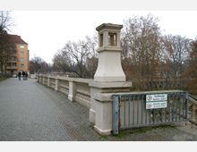 Fußweg entlang einer historischen Brücke mit Balustrade und einer Laternenhalterung in beigem Putzstil, umgeben von kahlen Bäumen und einem mehrstöckigen Wohngebäude im Hintergrund. Im Vordergrund ein Metallzaun mit Schildern, die Hunde und Ballspielen verbieten.