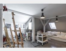 Helles Atelier unter einem Dach mit schrägen Wänden, Holzböden und großen Fenstern. Im Raum stehen Staffeleien, ein Sofa, Regale mit Büchern und Malutensilien sowie ein Rollwagen für Arbeitsmaterialien.