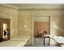 Innenansicht eines renovierungsbedürftigen Hauses mit roh verputzten, fleckigen Wänden und mehreren Türdurchgängen in angrenzende Räume. Im Vordergrund stehen provisorische Tische und Arbeitsplatten, im Hintergrund ein kleiner Tisch mit Stühlen auf einem staubigen Boden mit Bauschutt.