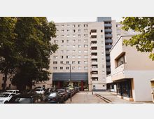 Wohnblock in einer städtischen Umgebung mit mehreren Etagen, Balkonen und einem Durchgang im Erdgeschoss. Im Vordergrund sind parkende Autos, ein Fußweg sowie Bäume zu sehen.