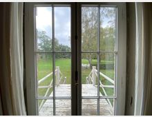 Blick durch ein großes, verglastes Doppelfenster auf eine hölzerne Veranda mit Geländer, die in eine grüne Wiese mit Bäumen und Gartenmöbeln im Hintergrund führt. Die Szene wird von Vorhängen an den Fensterseiten eingerahmt.