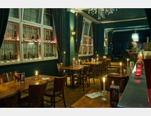 Restaurant im gehobenen Stil mit Holzmöbeln und dunkelgrünen Wänden, dekoriert mit Kerzen auf den Tischen und weinbestückten Regalen vor großen Fenstern; im Hintergrund ein Kronleuchter und eine Bar mit roten Teelichtern.