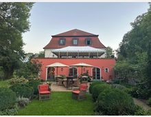 Ein zweistöckiges Haus mit rotem Dach und einer Terrasse mit Markise, umgeben von einem gepflegten Garten. Im Vordergrund stehen Sitzmöbel mit roten Polstern und Sonnenschirmen auf einer gepflasterten Fläche, umgeben von Hecken und Blumenbeeten.