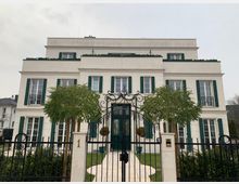 Dreistöckige Villa mit heller Fassade, grünen Fensterläden und dekorativen Balkonen, umgeben von einem schmiedeeisernen Zaun. Vor dem Eingangsbereich stehen kleine Bäume, und ein gepflasterter Weg führt zur Haustür.