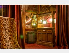 Ein historischer Innenraum mit einer hölzernen Theaterkasse, verziert mit Glasfenstern und geschwungenen Rahmen. Im Vordergrund ist ein Stuhl mit Leopardenmuster, umgeben von weinroten Vorhängen und Teppichboden.