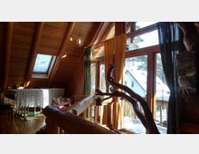 Innenraum eines Dachgeschosses aus Holz mit sichtbaren Balken und großen Fenstern, durch die Tageslicht einfällt. Vor den Fenstern hängen bunte Vorhänge, und draußen ist eine verschneite Landschaft mit einem Haus sichtbar; der Raum enthält einen Tisch mit Spitzen-Decke, Büchern und dekorative Elemente.