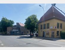 Straßenszene mit einer Kreuzung und zwei gegenüberliegenden Gebäuden: rechts ein gelbes Gebäude mit großen Fenstern und einem steilen Dach, links ein älteres Gebäude mit grauer Fassade und einem Laden mit blauen Schaufenstern. Im Hintergrund stehen Bäume, und die Straße ist von Lichtmasten gesäumt.