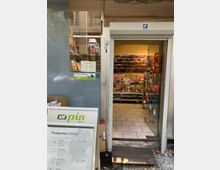 Außenansicht eines kleinen Ladens mit offener Eingangstür, hinter der ein Verkaufsraum mit Zeitschriftenregalen sichtbar ist. Neben der Tür steht ein Schild mit dem Logo von "PIN Mail" und Informationen zu Portopreisen.