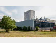 Flaches Gebäude mit blauer Glasfassade und wellenförmigem Dach, umgeben von einer Wiese und Bäumen im Vordergrund. Im Hintergrund steht ein mehrstöckiges Wohnhochhaus aus Beton.