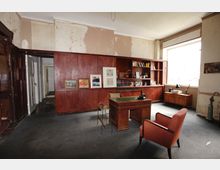 Geräumiges Büro mit großer dunkler Holzschrankwand und offenen Regalen, davor ein Holzschreibtisch und ein roter Polstersessel auf grauem Teppichboden. Helles Fenster an der rechten Seite, abblätternder Putz an den Wänden und eine Türöffnung links zu einem Flur.