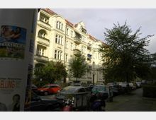 Straßenszene mit einem historischen Wohngebäude aus hellem Putz mit dekorativen Balkonen und roten Dächern. Im Vordergrund geparkte Autos und Mopeds sowie ein Baumgesäumter Gehweg; links eine Litfaßsäule mit Plakaten.