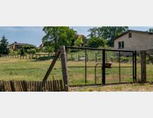 Eine ländliche Szene mit einem Maschendrahtzaun und einem Metalltor im Vordergrund. Dahinter befinden sich eine Wiese, mehrere Bäume und vereinzelt stehende Wohnhäuser mit einfacher Architektur.