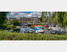 Kleiner Hafenbereich mit mehreren festgemachten Motorbooten und einem Hausboot, umgeben von Grünflächen und einem Weidenbaum im Vordergrund. Im Hintergrund sind moderne mehrstöckige Wohngebäude zu sehen, die sich entlang eines Gewässers erstrecken.