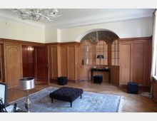 Ein Wohnzimmer mit hellen Holzwänden und verziertem Holzpaneel-Design. Der Raum enthält einen kleinen Sitzhocker, einen Teppich, eine Lampe, sowie eine dekorative Nische mit Glastüren und floralen Mosaiken oberhalb.