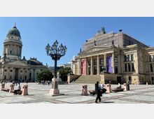 Das Bild zeigt den Gendarmenmarkt in Berlin mit dem Konzerthaus Berlin und dem Französischen Dom im Hintergrund. Der Platz ist gepflastert und mit Sitzbänken sowie einer Laterne ausgestattet, umgeben von historischen Gebäuden und besetzt von Spaziergängern.