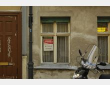 Eine alte Hausfassade mit einem leicht beschädigten Fensterrahmen, hinter dem weiße Gardinen hängen. Am Fenster ist ein Schild mit der Aufschrift „Coca-Cola Mittwoch Ruhetag“ angebracht. Links befindet sich eine hölzerne braune Tür mit Graffiti, und im Vordergrund steht ein Motorrad mit Windschutzscheibe.