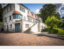 Weißes zweistöckiges Gebäude im Landhausstil mit roten Ziegeldächern, grünen Fensterläden und Natursteinelementen. Vor dem Gebäude befindet sich eine gepflasterte Auffahrt, eine Außentreppe und ein kleiner Garten mit Bäumen und Rasen.