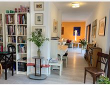 Wohnraum mit hellem Holzfußboden, Bücherregalen und einem weißen Klavier im Vordergrund. Im Hintergrund befindet sich ein Sitzbereich mit gelber Wand, einem Sessel, Tisch und Stehlampe, umgeben von Gemälden und Dekorationen.