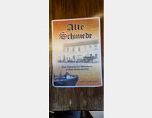 Flyer auf einer Holzoberfläche mit der Aufschrift „Alte Schmiede“ und einem historischen Foto eines zweigeschossigen Gasthauses. Darunter steht „Das historische Wirtshaus in Bad Liebenwerda“ sowie eine Telefon­nummer.