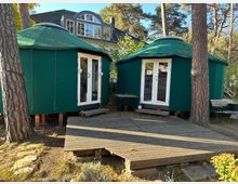 Zwei grüne, rundliche Zelte mit weißen Doppeltüren stehen auf einer Holzplattform inmitten eines bewaldeten Außenbereichs. Im Hintergrund ist ein Haus mit großen Fenstern und Dachgauben zu sehen.