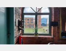 Innenansicht eines Backsteingebäudes mit Blick durch ein großes, sprossenverglastes Fenster auf einen Innenhof, umgeben von weiteren Backsteingebäuden und einem Gebäude mit Glasfassade. An der Wand neben dem Fenster hängt ein altes Telefon, darunter ein Feuerlöscher.