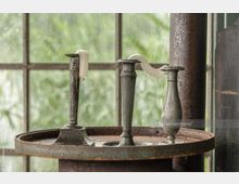 Drei metallene Kerzenhalter mit geschmolzenem, herunterlaufendem Wachs stehen auf einer rostigen, runden Metallplatte vor einem großen Fenster mit regennassen Scheiben und Blick auf grüne Vegetation.