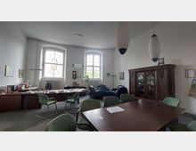 Ein Büroraum mit hohen Decken, zwei großen Fenstern und einer Mischung aus modernen und antiken Möbeln. Ein großer Besprechungstisch mit grünen Stühlen steht im Vordergrund, während im Hintergrund ein Schreibtisch und ein reich verzierter Holzschrank zu sehen sind.