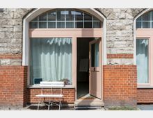 Eine Fassade aus roten und grauen Ziegelsteinen mit einer großen Fensterfront, die mit pastellfarbigen Holzrahmen eingefasst ist. Vor dem Fenster steht eine weiße Holzbank, die offene Tür führt in einen Raum mit Vorhängen und einem Flipchart.