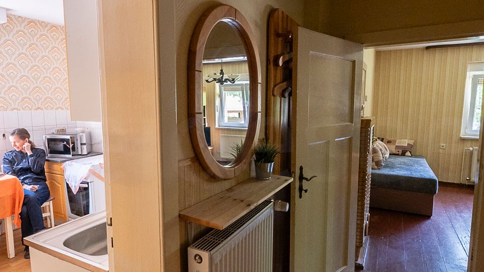 Schmaler Wohnungsflur mit Parkettboden, Wandheizkörper und runden Wandspiegel über einer kleinen Ablage; offene Türen führen links in eine Küche und geradeaus in ein Schlafzimmer mit Bett und Fenster.