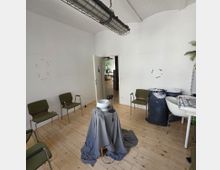 Ein minimalistischer Raum mit weißen Wänden, hellem Holzboden und einem offenen Türdurchgang im Hintergrund. Im Raum stehen grüne Stühle, ein Tisch mit Zeitungen, große Müllsäcke mit geschreddertem Papier und ein kleiner Tisch in der Mitte, bedeckt mit einem grauen Tuch, auf dem sich ein Keramikgegenstand und ein Mikrofon befinden.