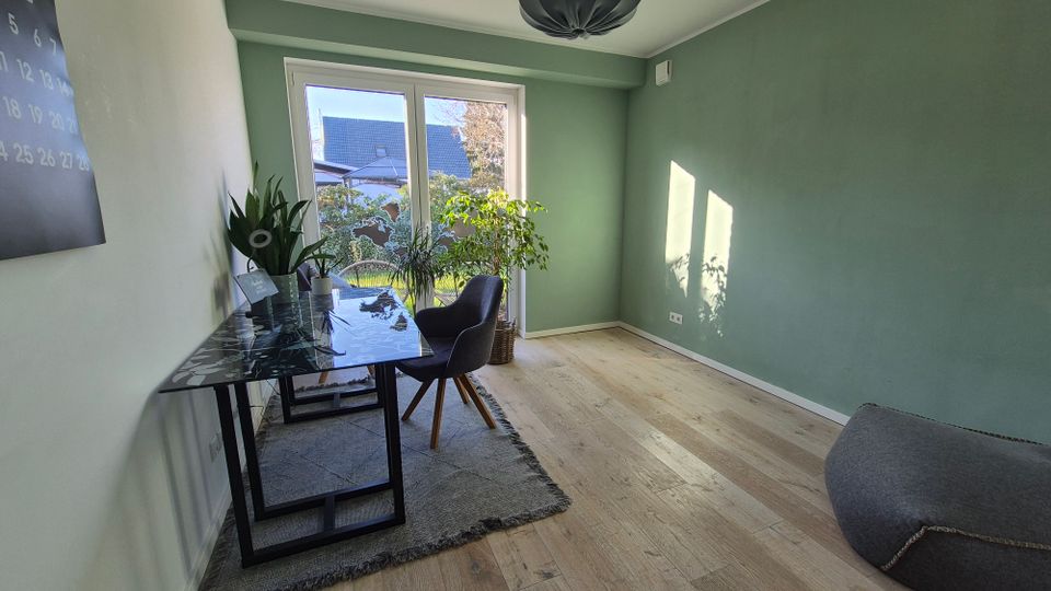 Minimalistisch eingerichtetes Arbeitszimmer mit grün gestrichenen Wänden, Holzfußboden und großem Fenster, durch das Tageslicht hereinfällt. Ein Schreibtisch aus Glas mit Pflanzen darauf und ein gepolsterter Stuhl stehen auf einem Teppich; rechts ein großer Sitzsack.