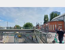 Fußgängerbrücke mit einem Geländer, unter der eine Straße verläuft. Rechts im Bild ein Backsteingebäude mit Rundbogenfenstern sowie Fahrräder und ein Gehweg, im Hintergrund ein moderner Bürokomplex mit der Aufschrift „BASF“ und eine Kirchturmspitze.