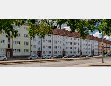 Straßenszene mit einer mehrstöckigen Wohnhausreihe im Hintergrund, die verputzt und in hellen Farben gestrichen ist, mit roten Satteldächern. Im Vordergrund befinden sich parkende Autos, Straßenbahnschienen und ein Verkehrsschild, während Bäume den oberen Bildrand säumen.