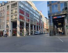 Straßenszene mit einem modernen Gebäude namens "Dussmann-Haus" mit großen Glasfassaden, roten Schildern und mehreren Geschäften im Erdgeschoss, darunter ein Vodafone-Shop. Im Hintergrund weitere Büro- und Geschäftsgebäude, rechts eine überdachte Passage, und auf der Straße geparkte Autos sowie Fußgänger.