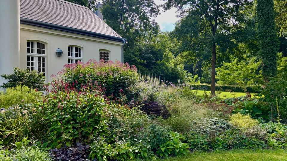 Heller Pavillon mit Schieferdach und Sprossenfenstern am Rand eines üppig bepflanzten Gartens mit Staudenbeeten und rosa Blüten. Daneben erstreckt sich eine Rasenfläche mit hohen Bäumen und dichtem Grün im Hintergrund.