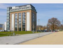 Modernes Mehrfamilienhaus mit Backstein- und weißen Fassadenelementen, großen Fenstern und einem Flachdach, umgeben von einem gepflasterten Gehweg, Grünflächen und parkenden Autos. Im Hintergrund stehen Bäume und weitere Gebäude.
