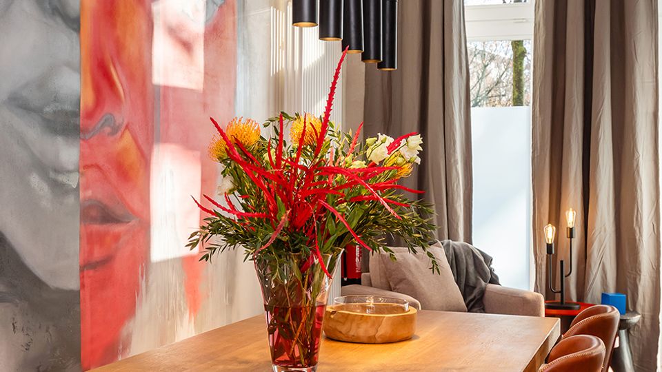 Moderner Essbereich mit langem Holztisch und braunen Lederstühlen, darüber eine Reihe schwarzer Pendelleuchten. An der Wand hängt ein großformatiges Wandbild, daneben bodentiefe Fenster mit grauen Vorhängen und Tageslicht.